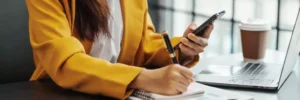 mulher fazendo anotações e vendo algo em celular, sentada à mesa de um escritório