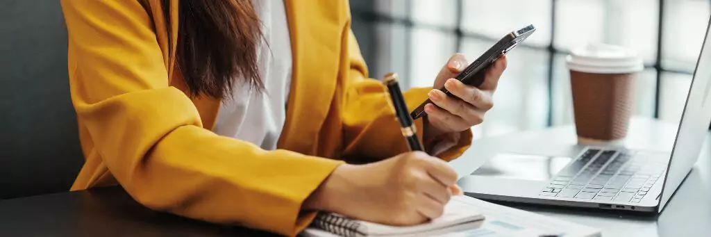 mulher fazendo anotações e vendo algo em celular, sentada à mesa de um escritório