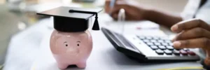 pessoa fazendo contas em calculadora com cofre de porquinho ao lado, com capelo na cabeça