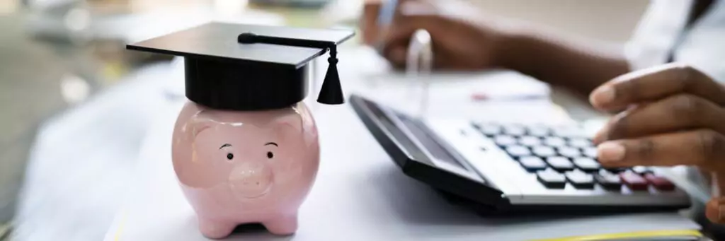 pessoa fazendo contas em calculadora com cofre de porquinho ao lado, com capelo na cabeça